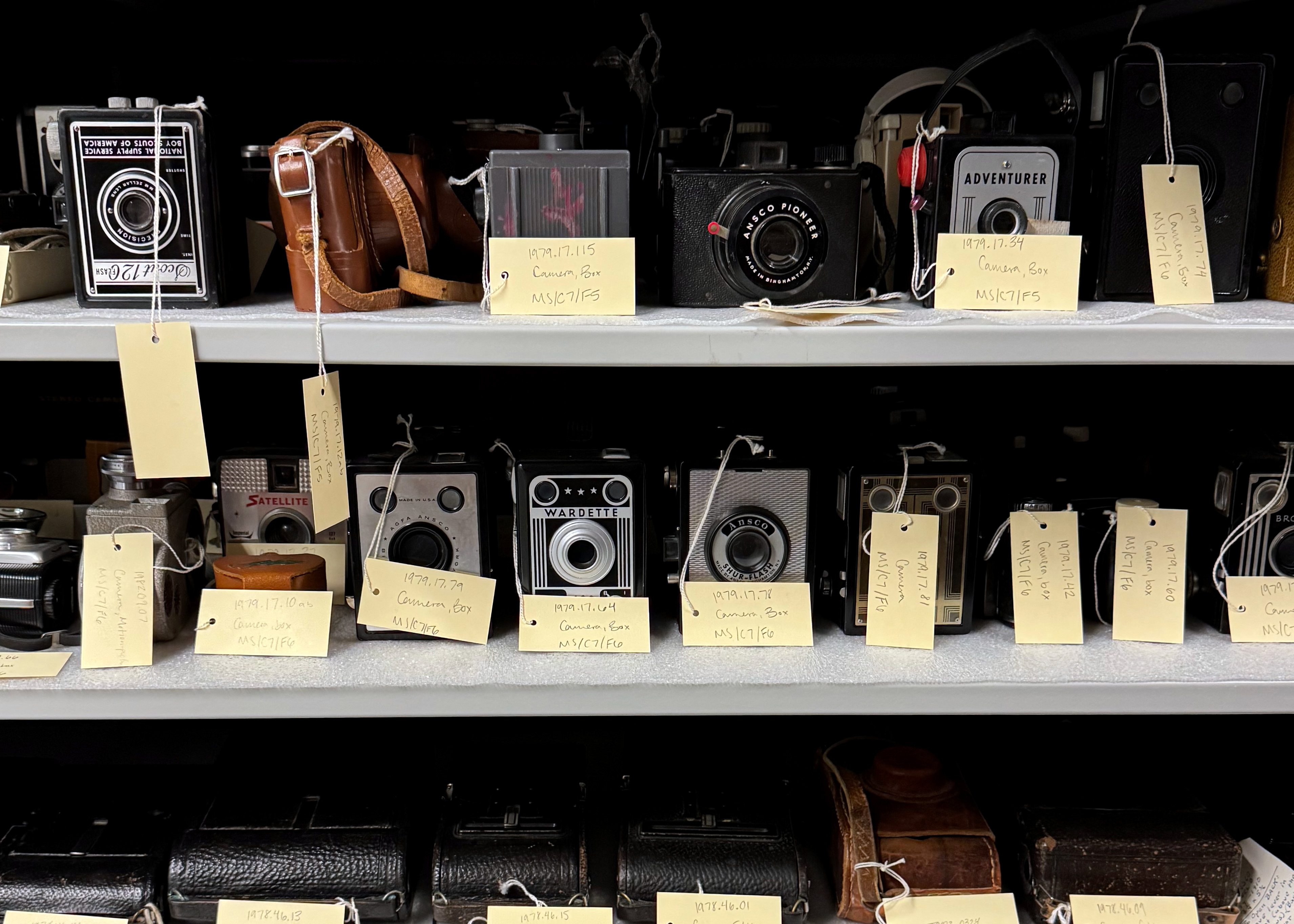 a collection of vintage and historical cameras are on shelves with collection tags on them